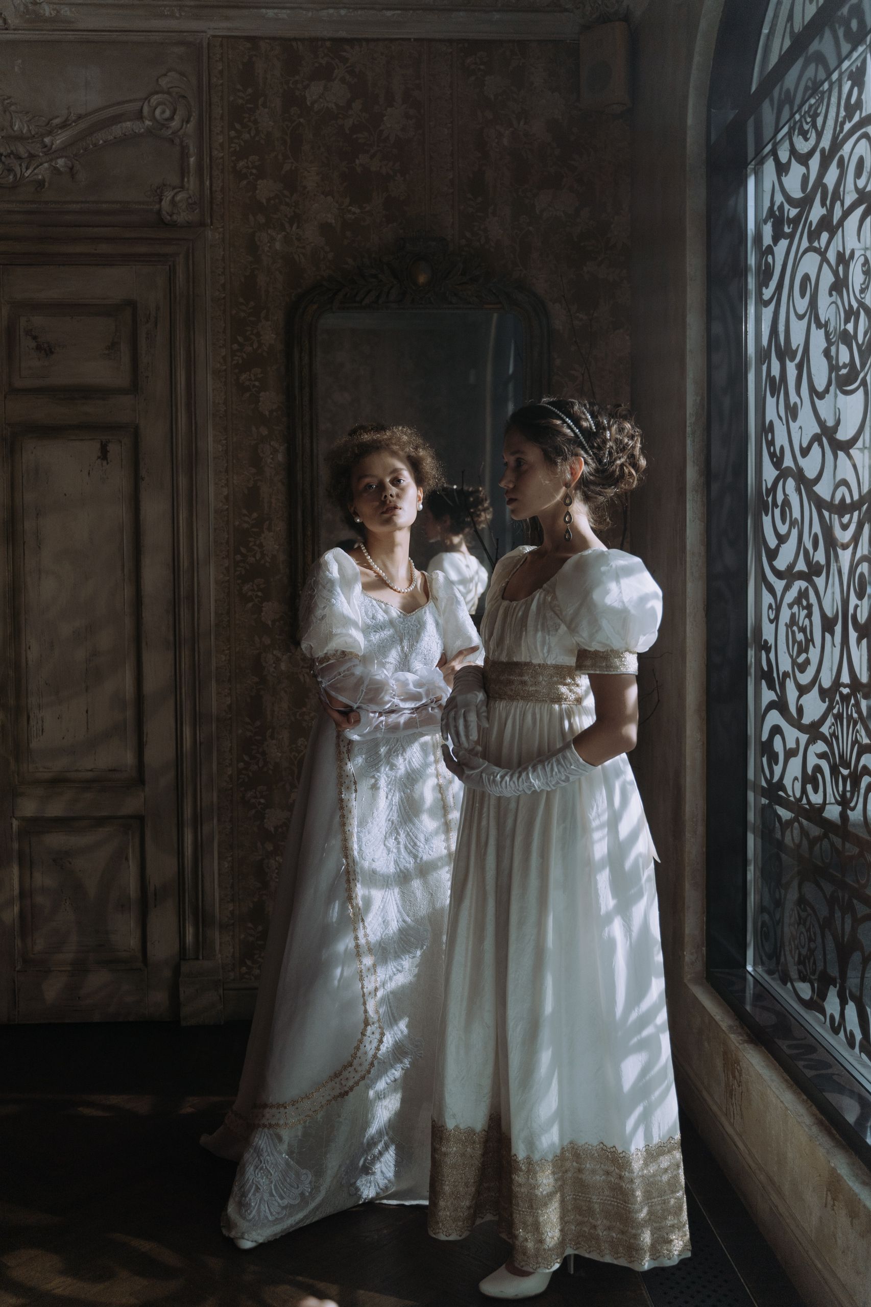 Two women in Victorian-style dresses stand elegantly in a dimly lit, ornate room.