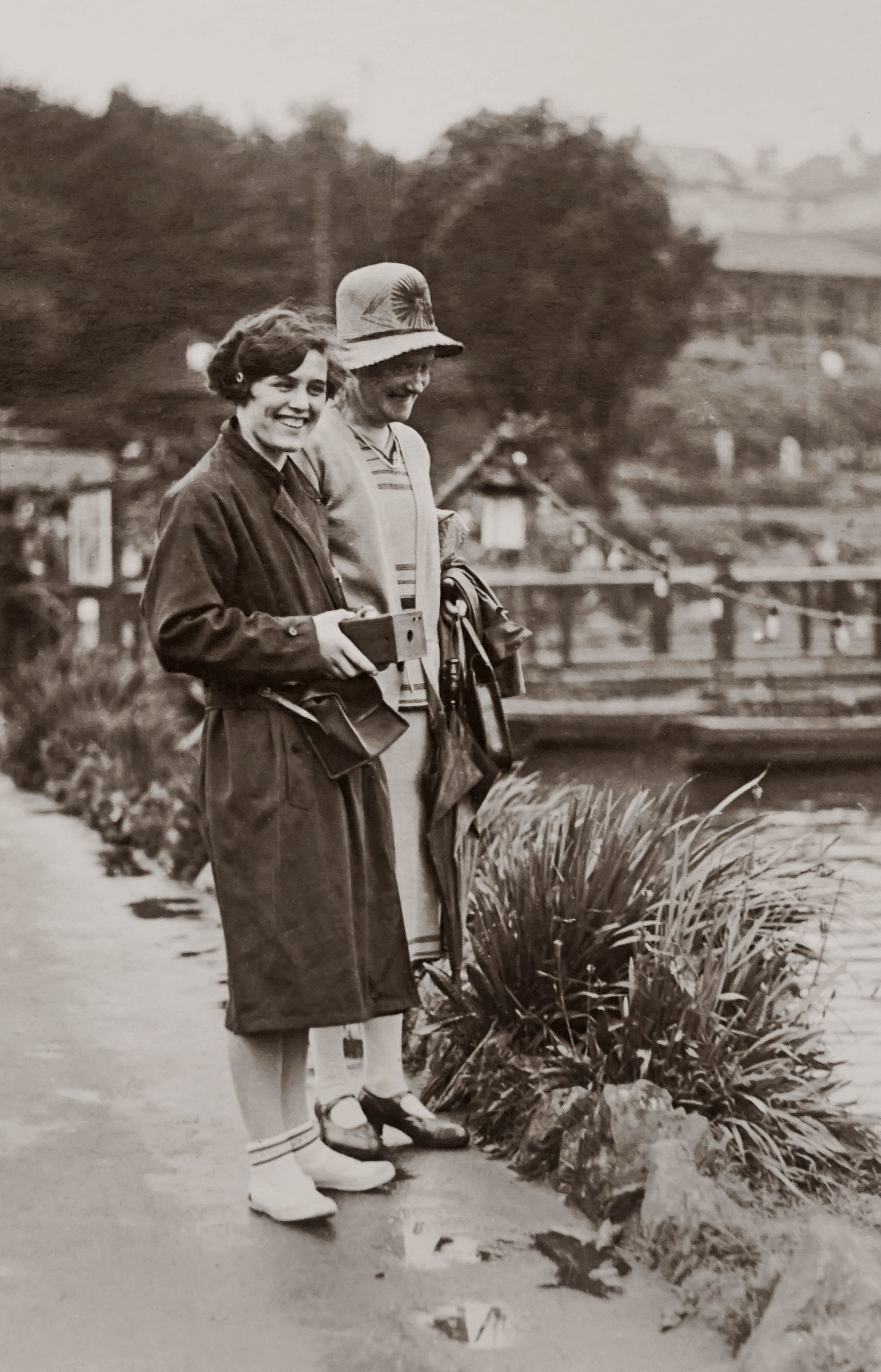 Two women in vintage attire smiling by a riverside path, a classic monochrome photograph.